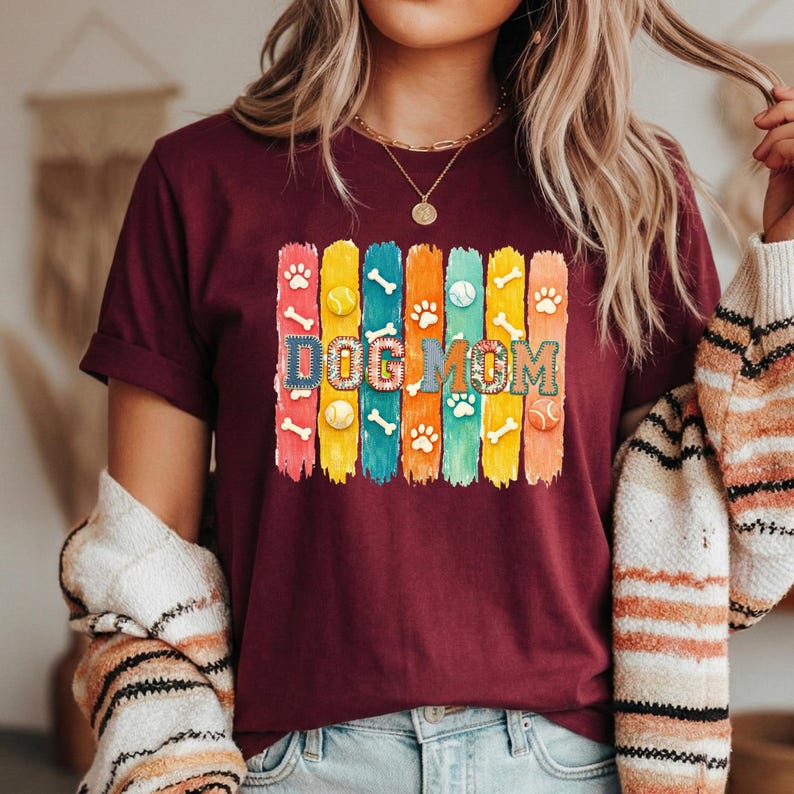 Colorful dog mom graphic with vertical painted stripes, paw prints, bones, and tennis balls in a playful patchwork style design for shirts and sublimation.