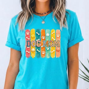 Colorful dog mom graphic with vertical painted stripes, paw prints, bones, and tennis balls in a playful patchwork style design for shirts and sublimation.