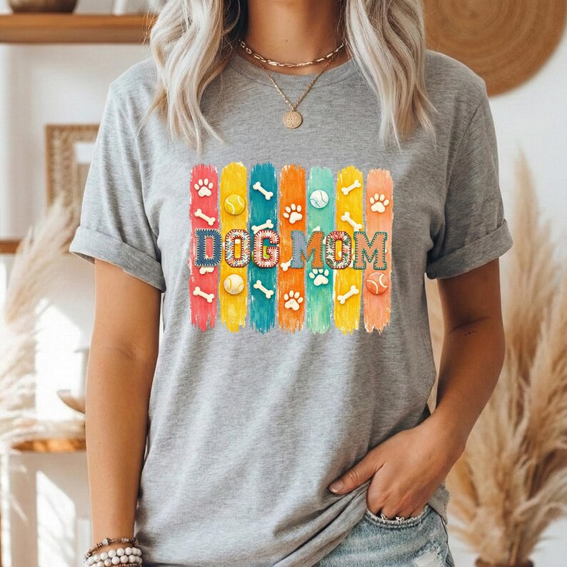 Colorful dog mom graphic with vertical painted stripes, paw prints, bones, and tennis balls in a playful patchwork style design for shirts and sublimation.