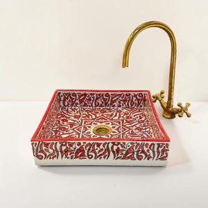 Può includere: Un lavabo quadrato in ceramica con base bianca e intricati motivi floreali e geometrici rossi dipinti a mano. Il lavabo include uno scarico centrale in ottone ed è abbinato a un rubinetto in ottone alto e curvo con due manopole a croce.