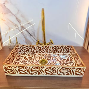 Op de afbeelding: Een rechthoekige keramische wastafel met een witte basis en bruine, handgeschilderde bloem- en geometrische patronen. De wastafel is voorzien van een centrale messing afvoer en een hoge, gebogen messing kraan met twee handgrepen.