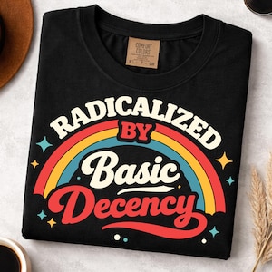 May include: Black t-shirt with the phrase "RADICALIZED BY Basic Decency" in a retro rainbow design. The shirt has a crew neck and a small tag on the upper back. The design includes stars and a rainbow.