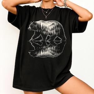 Puede incluir: Camiseta negra con un diseño en escala de grises de un oso caminando en el agua, reflejando un bosque y montañas. El contorno del oso es detallado, y el diseño tiene un tema de naturaleza.