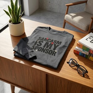 以下が含まれることがあります： ダークグレーの長袖Tシャツ。前面中央に黒い大文字で「SARCASM IS MY SPONSOR」というテキストがプリントされており、小さな赤いひし形と白い四芒星のアイコンがアクセントとしてあしらわれています。