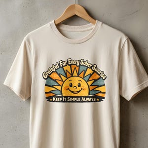 以下が含まれることがあります： オレンジと青の光線を持つ笑顔の太陽のグラフィックが描かれたクリーム色の半袖Tシャツ。太陽の上には「Grateful For Every Sober Sunrise」、太陽の下には2つの星と共に「KEEP IT SIMPLE ALWAYS」というテキストがプリントされています。