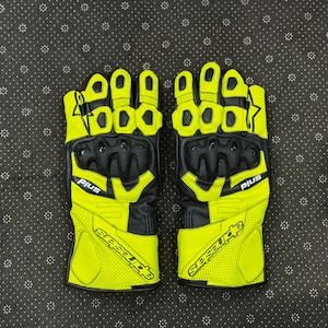 Könnte beinhalten: Paar neongelbe und schwarze Motorradhandschuhe. Die Handschuhe haben ein perforiertes Design auf den gelben Bereichen, mit schwarzer Polsterung an Knöcheln und Handfläche. Der Markenname "Alpinestars" ist auf dem Handgelenk aufgedruckt.
