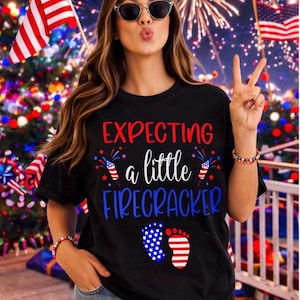 May include: Black t-shirt with the text "EXPECTING a little FIRECRACKER" in red and blue. The shirt features firecracker and footprint graphics. The background includes fireworks and American flags.
