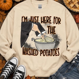 May include: A beige crewneck sweatshirt featuring a graphic of a hand in a black suit sleeve whisking mashed potatoes in a gray pot. The text reads, "I'M JUST HERE FOR THE MASHED POTATOES" in black and orange lettering with a gray splatter background.