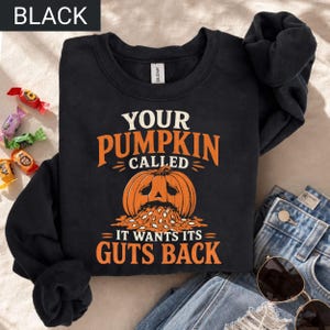 May include: A black long-sleeve sweatshirt featuring a graphic of a carved pumpkin with its seeds spilling out. The text reads: YOUR PUMPKIN CALLED IT WANTS ITS GUTS BACK. The design uses orange and cream lettering on the front of the garment.