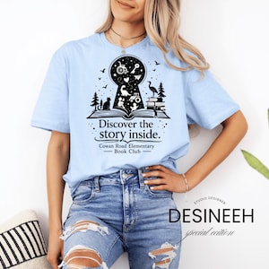 May include: Light blue t-shirt with a black graphic featuring a keyhole design with a book, cat, and other illustrations. Text reads "Discover the story inside." and "Cowan Road Elementary - Book Club."