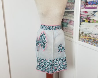 May include: A vintage blue and green floral apron with pink trim and a large pocket. The apron is on a dress form.