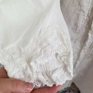 May include: White cotton blouse with a lace trim around the cuffs. The lace is a delicate eyelet pattern.