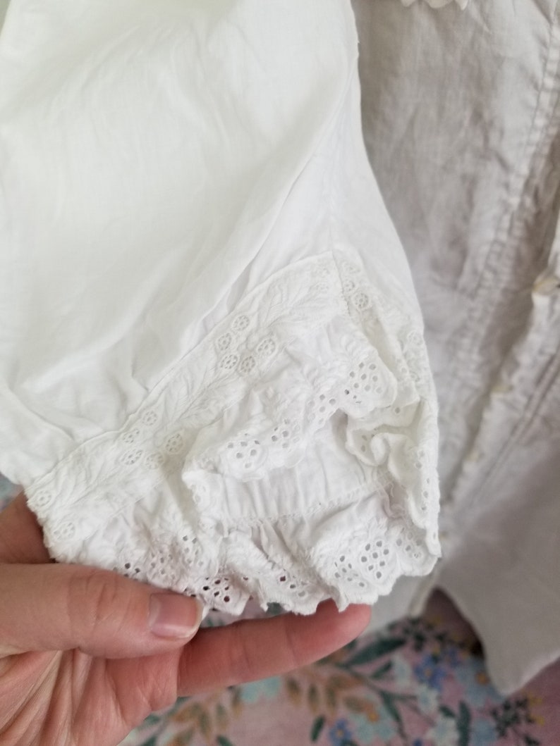May include: White cotton blouse with a lace trim around the cuffs. The lace is a delicate eyelet pattern.