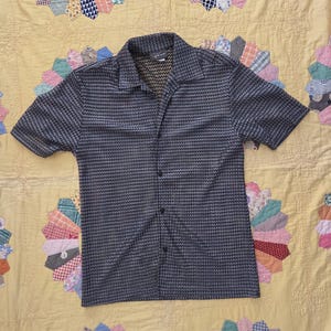 Può includere: Una camicia abbottonata a maniche corte con un motivo a quadri blu scuro e bianco. La camicia ha un colletto ed è realizzata in un tessuto leggero. La camicia è esposta su una trapunta gialla con motivi circolari colorati.