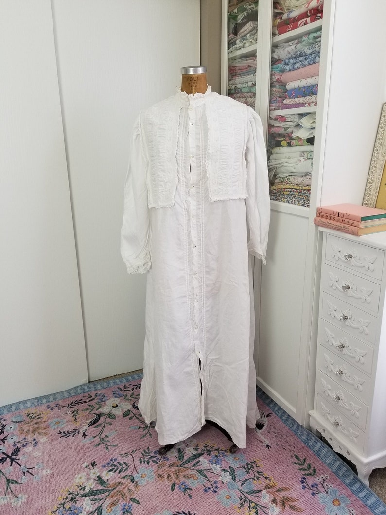 May include: A white cotton nightgown with a ruffled collar and long sleeves. The gown has a button-up front and is on a white mannequin.