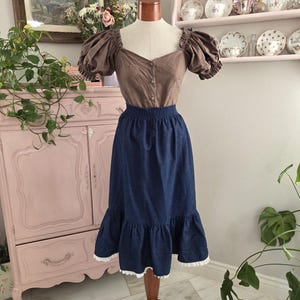 May include: A brown blouse with short, puffed sleeves and a button-down front is paired with a dark blue denim skirt. The skirt has a ruffled hem with white lace trim. The outfit is displayed on a mannequin.