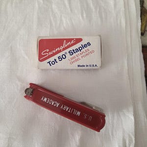 May include: A red Swingline stapler and a box of Swingline Tot 50 staples on a white cloth. The stapler has the text "U.S. MILITARY ACADEMY" printed on it. The staple box reads "1,000 STAPLES CHISEL POINTED" and "Made in U.S.A."