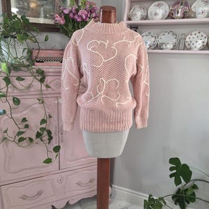 May include: A light pink sweater with a high neckline and long sleeves. The sweater features a white, heart-shaped design. The sweater is displayed on a mannequin with a wooden stand. The background includes a pink cabinet and decorative items.