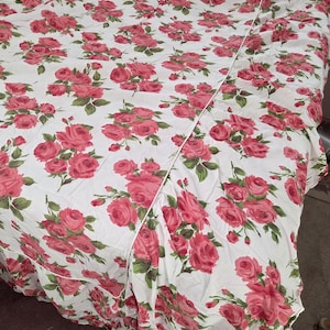 May include: White bedspread with a pattern of pink roses and green leaves. The floral design covers the entire surface of the bedspread. The bedspread has a ruffled edge and a seam down the middle.