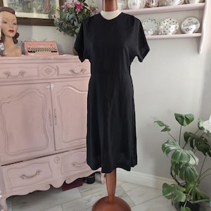 Könnte beinhalten: Ein schwarzes Kleid mit kurzen Ärmeln, präsentiert auf einer Holzpuppe. Das Kleid hat ein schlichtes, klassisches Design mit taillierter Taille und knielangem Saum. Die Puppe steht auf einem Holzständer mit rundem Sockel. Der Hintergrund umfasst einen rosa Schrank und Dekorationsartikel.