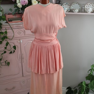 May include: A peach-coloured dress displayed on a mannequin, featuring short sleeves, a gathered waistline, and a pleated peplum detail that falls over a straight skirt. The garment has a simple round neckline and a smooth, solid-coloured fabric finish.