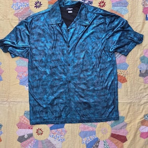 Può includere: Camicia turchese a maniche corte con bottoni e un motivo sottile. La camicia ha un colletto classico e una fodera interna nera. È appoggiata su una trapunta gialla vintage con disegni floreali colorati.