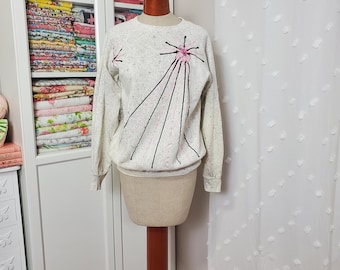 May include: A light gray sweatshirt with a speckled pattern, featuring a pink and black starburst design on the front. The long-sleeved top is displayed on a mannequin, showcasing its vintage style. The sweatshirt is a vintage piece.