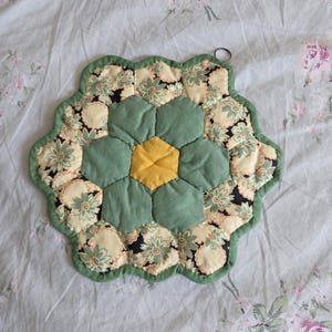 May include: A handmade, flower-shaped pot holder. The center is yellow, surrounded by green and cream-colored hexagonal fabric pieces with floral patterns. A green border and a small hanging loop complete the design. The pot holder is on a floral patterned background.
