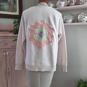 Pode incluir: Um moletom rosa claro com um design de patchwork floral nas costas. O patchwork apresenta formas geométricas rosa, verde e roxa. O moletom é exibido em um manequim.