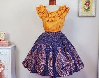 May include: A vintage-style skirt with a blue background and a gold and pink paisley pattern. The skirt is full and has a gathered waist. It is paired with a yellow ruffled blouse.