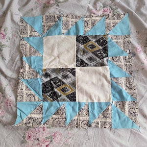 May include: A square quilt block featuring a central four-patch design with two solid off-white squares and two squares with a black, white, and gold geometric pattern. The border consists of light blue triangles and black and white floral squares.
