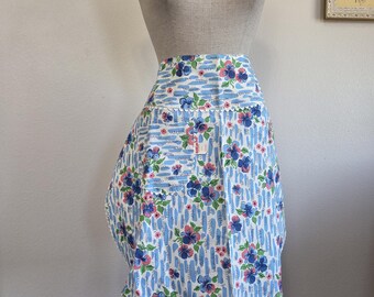 May include: A vintage-style apron displayed on a mannequin. The apron features a light blue background with vertical white stripes and a floral pattern in blue and pink. The apron has a scalloped edge.