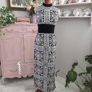 May include: A sleeveless black and white patterned dress with a high collar and a black waist belt. The dress is displayed on a mannequin. The pattern is a repeating floral design. The dress is floor length.