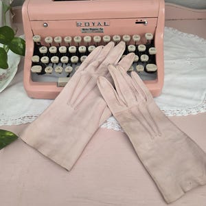 May include: A pair of pale pink leather gloves with stitched details, resting on a pink surface in front of a vintage pink Royal typewriter. The typewriter has white keys and the text "ROYAL" is visible.