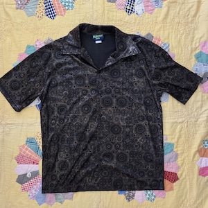 Può includere: Una camicia marrone scuro a maniche corte con un motivo geometrico. La camicia ha un colletto e uno scollo a V. L'etichetta recita "Plasso". La camicia è esposta su una trapunta gialla con motivi circolari colorati.