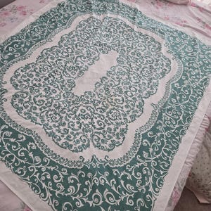 May include: A white and teal patterned textile with an ornate design. The pattern features swirling floral and scroll motifs, framed by a teal border. The textile is laid out on a bed with a floral patterned bedspread.