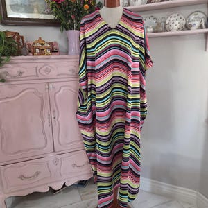 May include: A floor-length kaftan dress featuring a vibrant, multi-colored wavy stripe pattern in shades of black, bright pink, lime green, and light blue. The garment has a V-neckline, short sleeves, and a loose, flowing silhouette on a mannequin.
