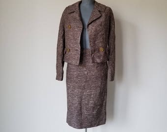 Pode incluir: Um conjunto de saia de tweed marrom com uma jaqueta curta. A jaqueta tem dois botões dourados e uma gola. A saia é de comprimento até o joelho e tem uma silhueta reta.