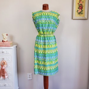 Puede incluir: Un vestido vintage sin mangas en tonos azules, verdes y amarillos, exhibido en un maniquí de madera. El vestido presenta un patrón geométrico y una cintura ceñida. El vestido llega a la rodilla.