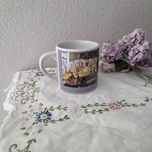 Könnte beinhalten: Weiße Keramik-Kaffeetasse mit Henkel, mit einem Gemälde von Menschen in einer Parkszene. Die Tasse hat einen lavendelfarbenen Rand und steht auf einer weißen, bestickten Tischdecke mit floralen Mustern. Ein Strauß lila Blumen befindet sich im Hintergrund.