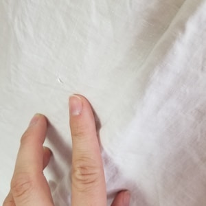 May include: Close-up of a white fabric surface with a small hole in the centre. A hand is touching the fabric.