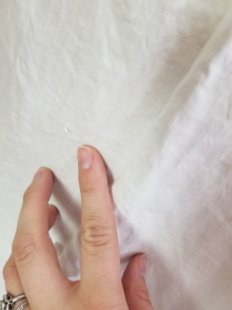 May include: Close-up of a white fabric surface with a small hole in the centre. A hand is touching the fabric.