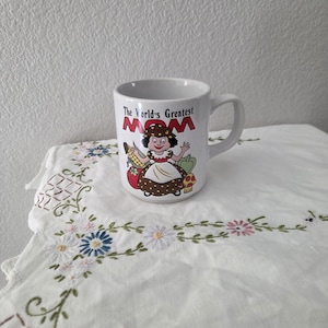 May include: White ceramic mug with the text "The World's Greatest Mom" in red and brown. The mug features a cartoon illustration of a woman holding vegetables. The mug is on a white embroidered tablecloth.