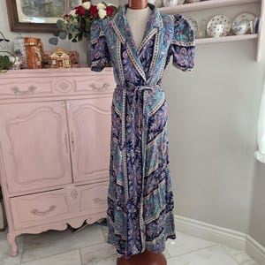 May include: A floor-length wrap dress featuring a dark blue, light blue, and purple paisley pattern. The garment has short puff sleeves, a notched collar, and a matching fabric tie belt at the waist, displayed on a mannequin.