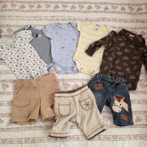 May include: A collection of baby clothes, including onesies and shorts, in various colors and patterns. The onesies feature stripes, prints, and gingham patterns. Shorts are khaki, tan, and denim, some with embroidered details.