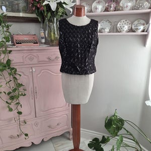 May include: A sleeveless black top featuring an all-over sequined chevron pattern. The garment has a round neckline and a straight hem, displayed on a beige fabric dress form with a wooden neck piece and a wooden stand base.