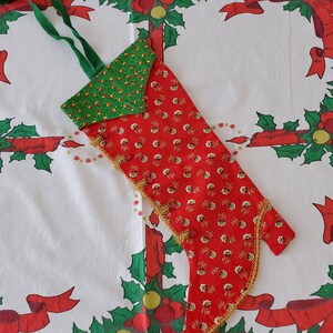 May include: A red and green Christmas stocking with gold trim and a green and yellow patterned top. The stocking is lying on a white tablecloth with a red and green holly pattern.