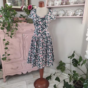 May include: A vintage-style dress on a mannequin. The dress features a dark background with a floral pattern of pink and white flowers. It has a fitted bodice and a flared skirt. The dress is displayed in a room with a pink cabinet and decorative plates.