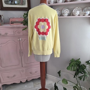 May include: A light yellow sweatshirt with a colorful floral design on the back. The design features red, white, and purple petals with a light blue base. The sweatshirt is displayed on a wooden dress form.