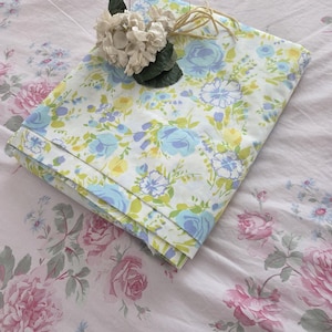 Puede incluir: Juego de sábanas blancas dobladas con un estampado floral en azul, amarillo y verde. Una flor decorativa con hojas verdes y cuerda está encima del juego de sábanas dobladas. El fondo es una sábana con estampado floral.
